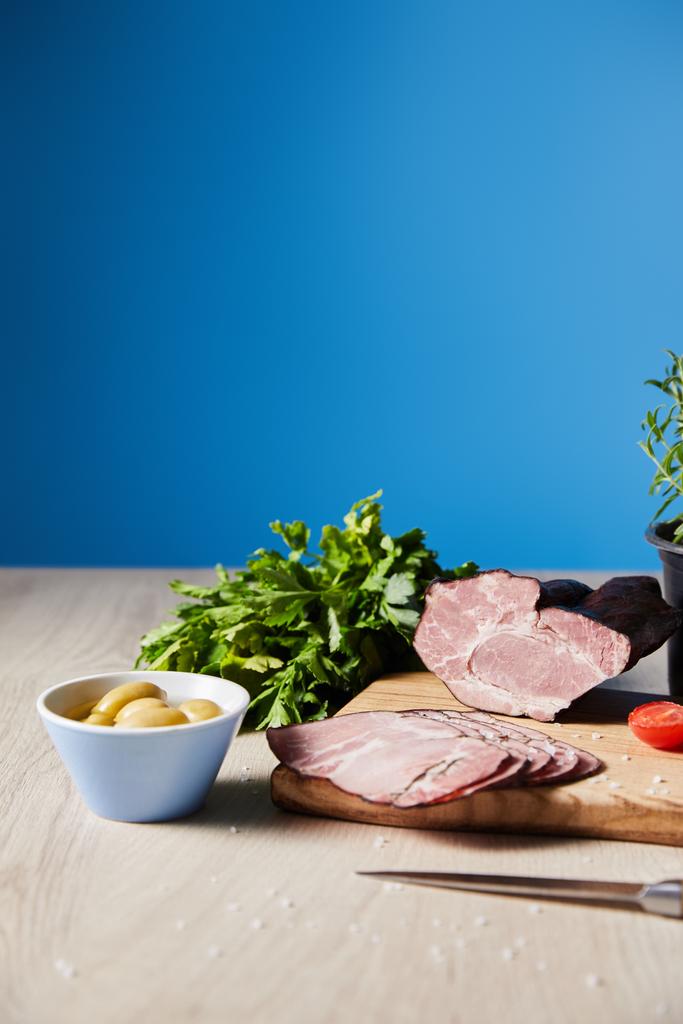 foco seletivo de presunto saboroso na placa de corte com faca, salsa, azeitonas na mesa de madeira no fundo azul
 - Foto, Imagem