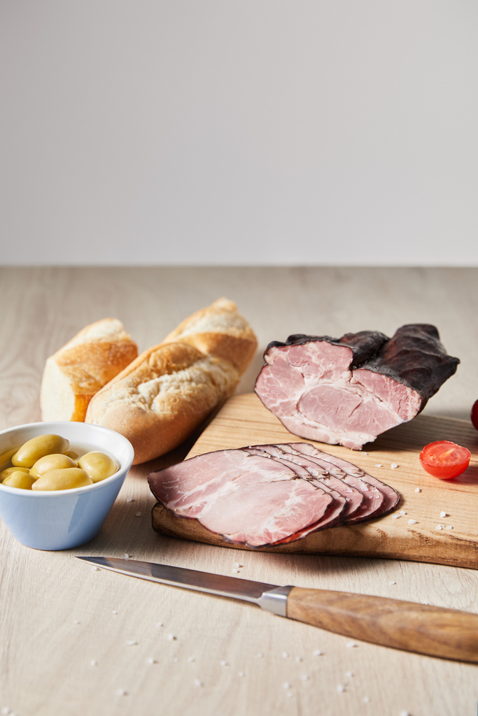 tasty ham on cutting board with knife, cherry tomato, olives and baguette on wooden table isolated on grey - Photo, Image