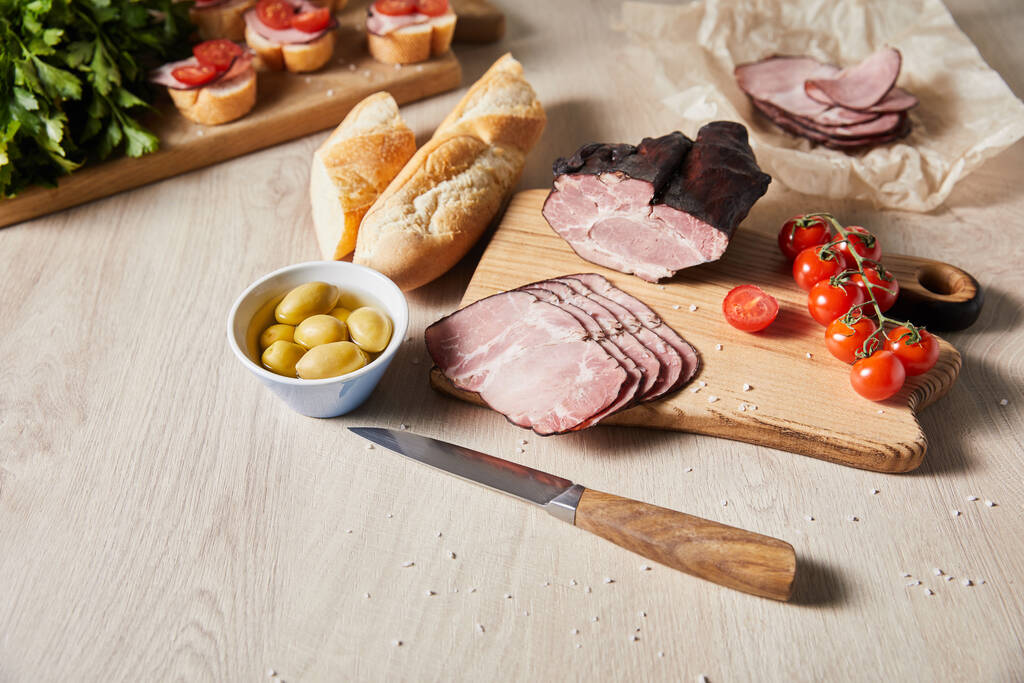 selectieve focus van smakelijke ham op snijplank met mes, peterselie, kerstomaten, olijven en stokbrood bij canape op houten tafel - Foto, afbeelding