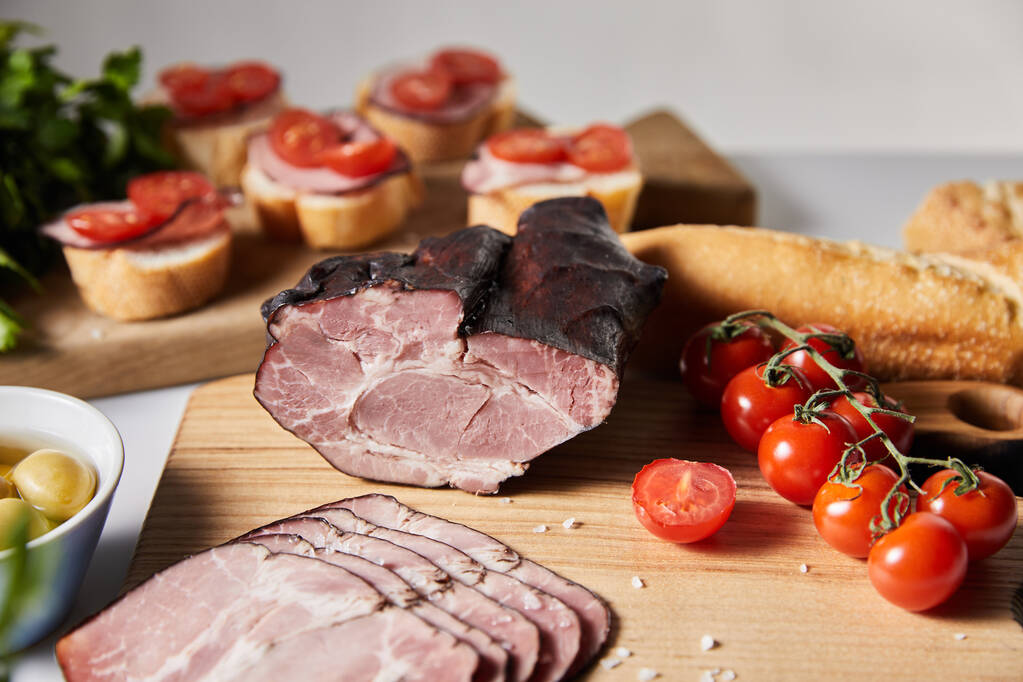 selektywny nacisk smacznej szynki na deskę do krojenia z pietruszką, pomidorami wiśniowymi, oliwkami i bagietką w pobliżu kanapek izolowanych na szaro - Zdjęcie, obraz