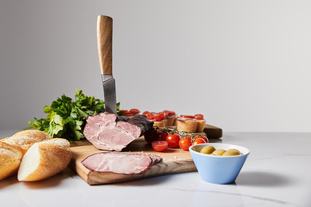selective focus of knife in tasty ham on cutting board with parsley, cherry tomatoes, olives and baguette on white surface isolated on grey - Photo, Image