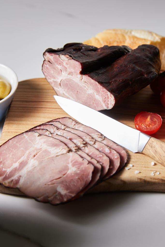 selective focus of tasty ham on cutting board with knife, cherry tomato and baguette on white surface - Photo, Image