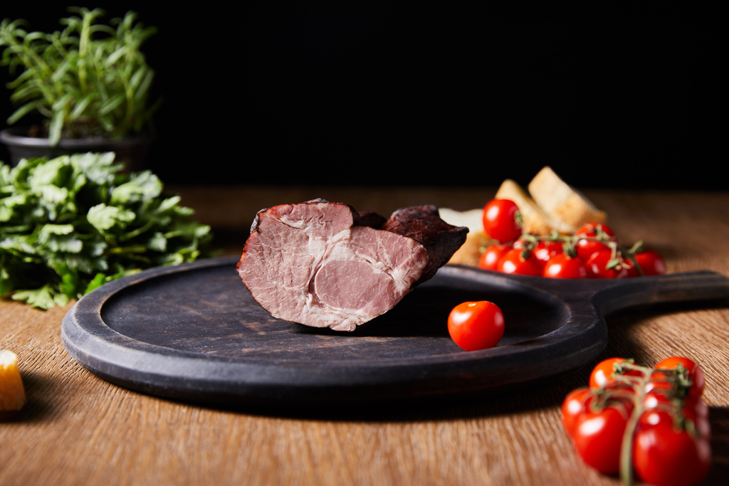 selective focus of tasty ham on board near parsley, cherry tomatoes and baguette on wooden table isolated on black - Photo, Image