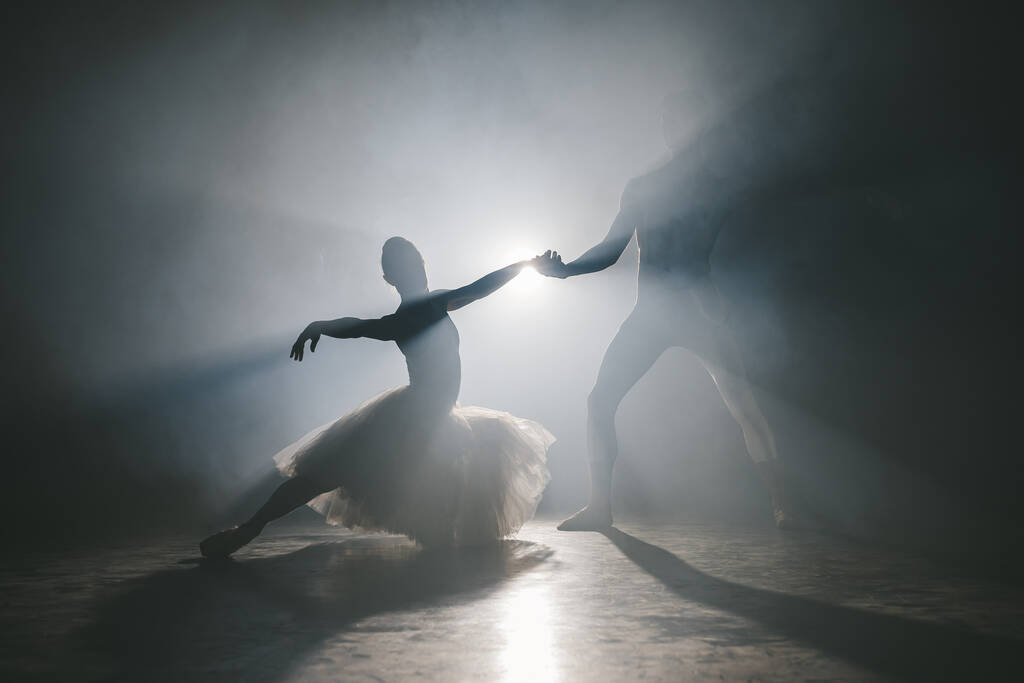 Η χαριτωμένη μπαλαρίνα και ο σύντροφός της χορεύουν στοιχεία κλασικού ή σύγχρονου μπαλέτου στο σκοτάδι με φόντο το φως του προβολέα. Ζευγάρι σε καπνό σε μαύρο φόντο. Καλλιτεχνική έννοια. - Φωτογραφία, εικόνα