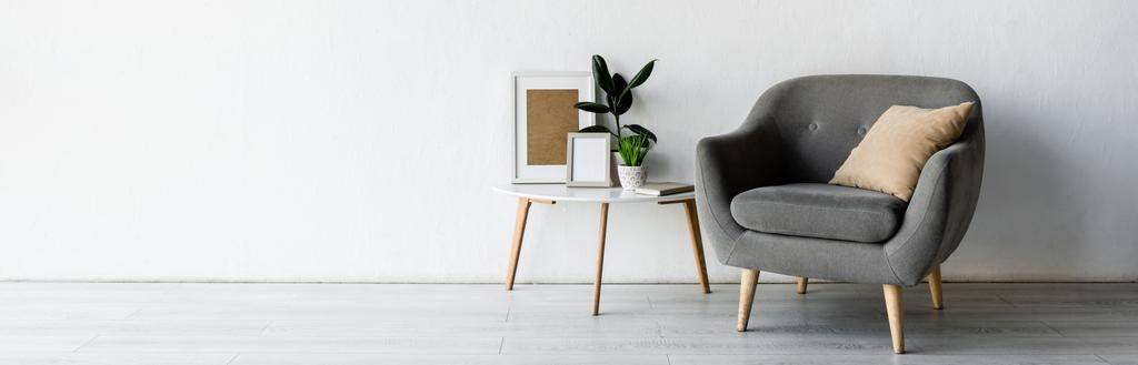 panorámás tájolás kényelmes fotel mellett dohányzóasztal zöld növények és keretek a modern nappaliban  - Fotó, kép