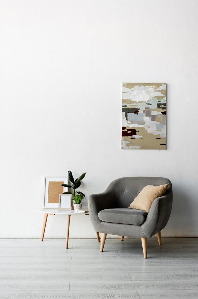 сіре крісло біля журнального столика з рамами і зеленими рослинами біля картини на стіні в сучасній вітальні
  - Фото, зображення