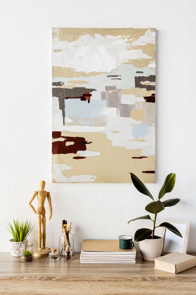 картина на стіні біля столу з дерев'яним манекеном, рослиною, блокнотами, книгою, чашкою, вазою з пшеницею та рамою
  - Фото, зображення
