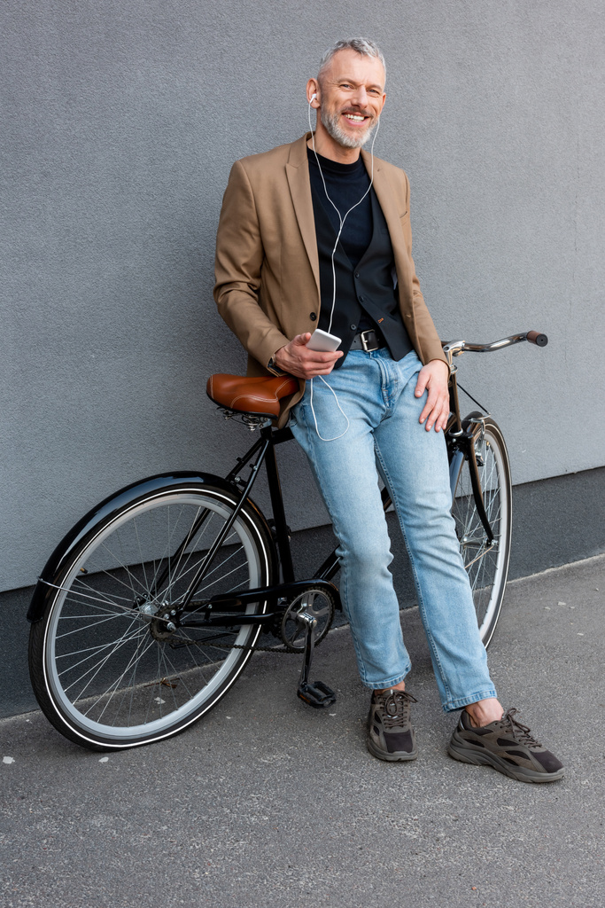 fröhlicher Geschäftsmann mit Kopfhörer, der Musik hört, während er sein Smartphone in der Hand hält und draußen neben dem Fahrrad steht  - Foto, Bild