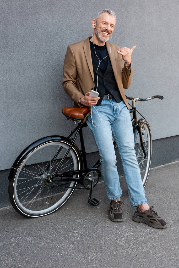 χαρούμενα επιχειρηματίας σε ακουστικά ακούγοντας μουσική, ενώ δείχνει κλήση μου χειρονομία και κρατώντας smartphone κοντά στο ποδήλατο έξω  - Φωτογραφία, εικόνα