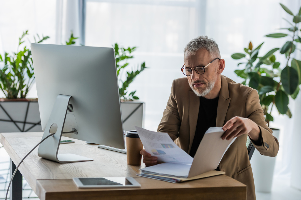selective focus of bearded businessman in glasses looking at document near coffee to go and computer monitor in office  - Photo, Image