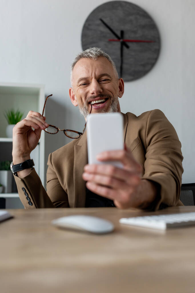 foyer sélectif d'homme d'affaires heureux tenant des lunettes et regardant smartphone dans le bureau
  - Photo, image