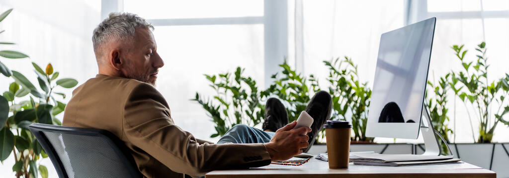 πανοραμική λήψη του επιχειρηματία που κάθεται κοντά στην οθόνη του υπολογιστή με λευκή οθόνη και κρατώντας smartphone  - Φωτογραφία, εικόνα
