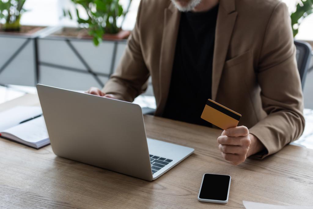 vista cortada de empresário segurando cartão de crédito perto de laptop e smartphone com tela em branco
  - Foto, Imagem