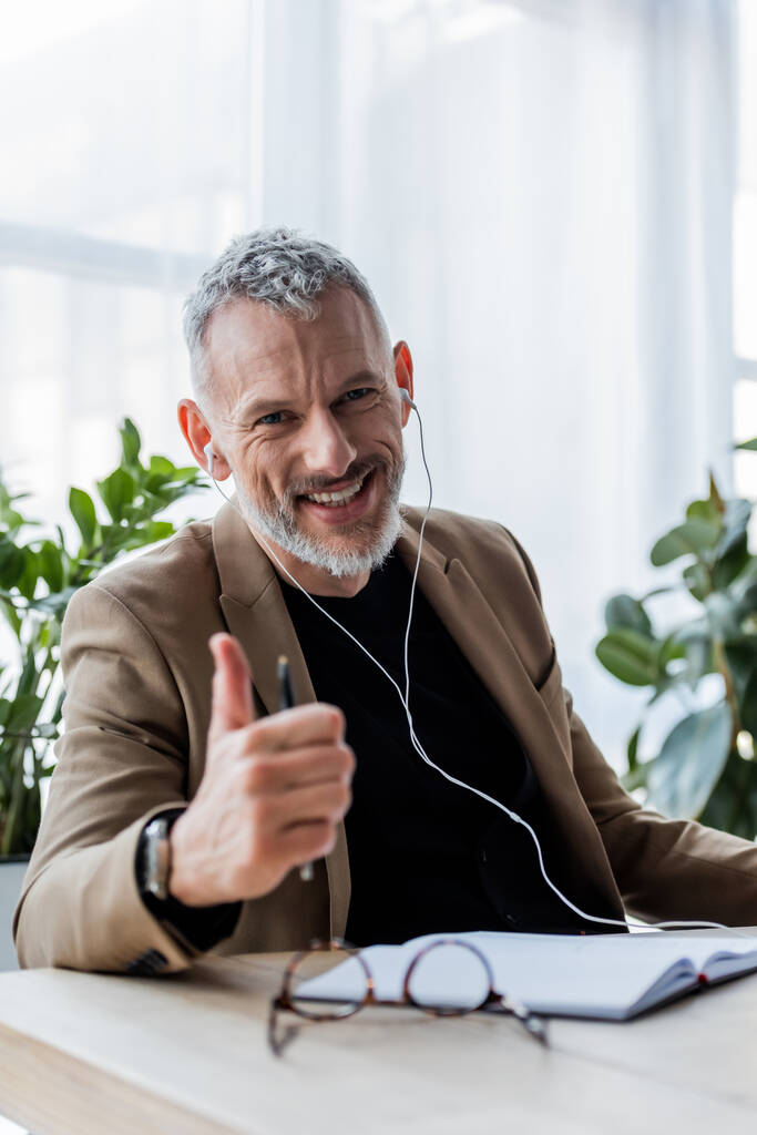 selective focus of happy businessman in earphones showing thumb up in office  - Photo, Image