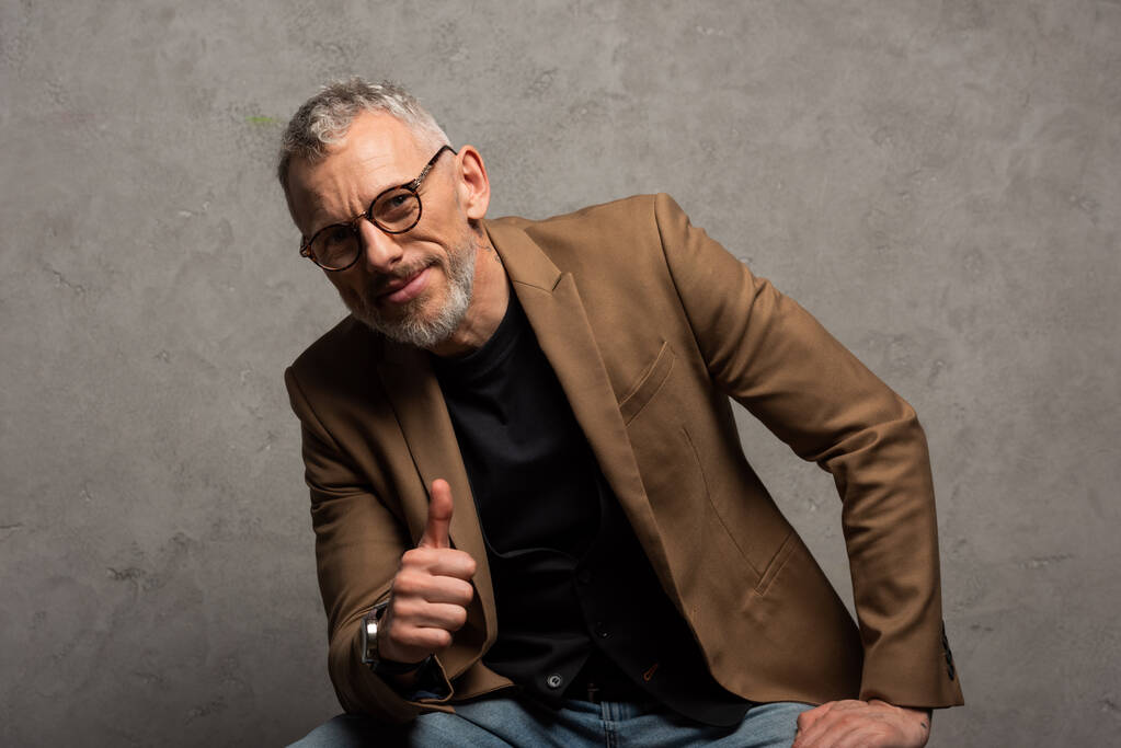 happy businessman in glasses looking at camera and showing thumb up on grey  - Photo, Image