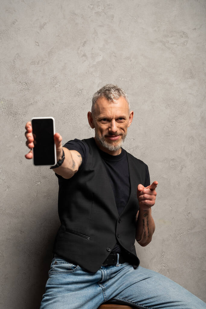 γενειοφόρος άνδρας κρατώντας smartphone με κενή οθόνη και δείχνοντας με το δάχτυλο σε γκρι  - Φωτογραφία, εικόνα