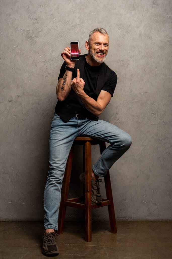 šťastný muž s tetováním ukazuje prstem na smartphone s obchodními kurzy na šedé  - Fotografie, Obrázek