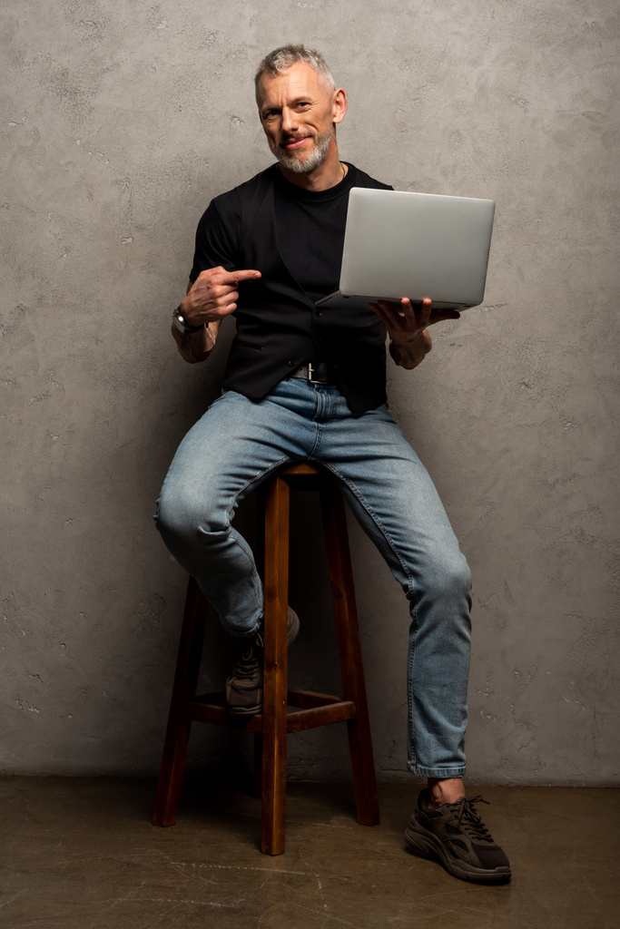 happy man pointing with finger at laptop and looking at camera on grey - Photo, Image