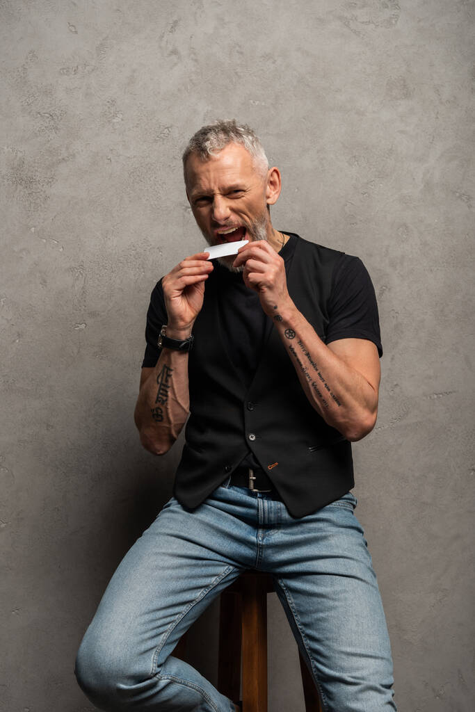 schöner Mann mit Tätowierungen beißt Blanko-Karte auf grau  - Foto, Bild