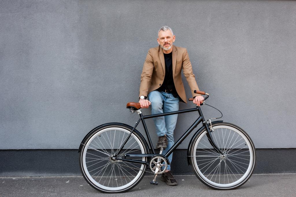 όμορφος επιχειρηματίας με σακάκι και τζιν στέκεται κοντά στο ποδήλατο έξω  - Φωτογραφία, εικόνα
