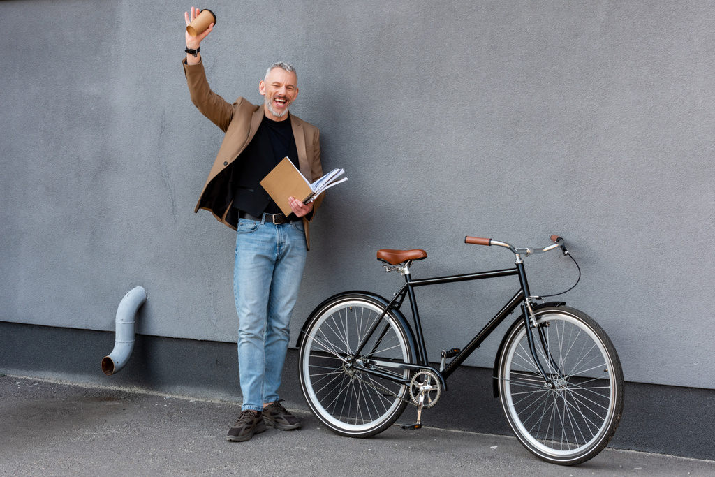 szczęśliwy biznesmen machając ręką trzymając papierową filiżankę i folder i stojąc w pobliżu roweru  - Zdjęcie, obraz