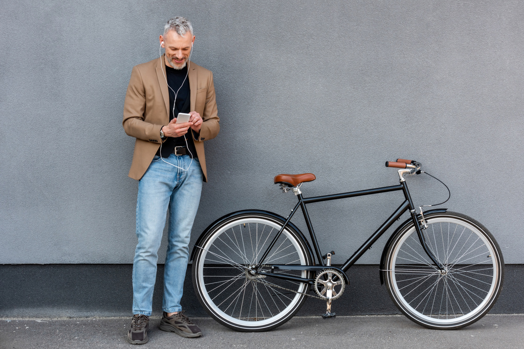 feliz hombre de negocios en auriculares escuchando música mientras sostiene el teléfono inteligente cerca de la bicicleta fuera
  - Foto, Imagen