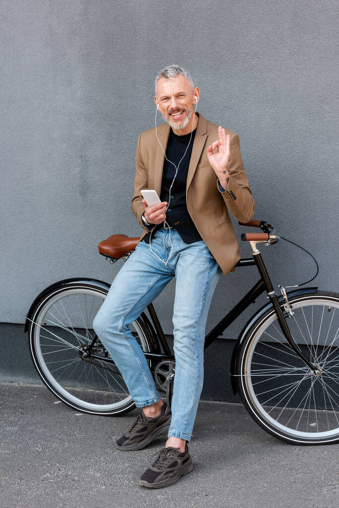 cheerful businessman in earphones listening music while showing ok sign and holding smartphone near bicycle outside  - Photo, Image