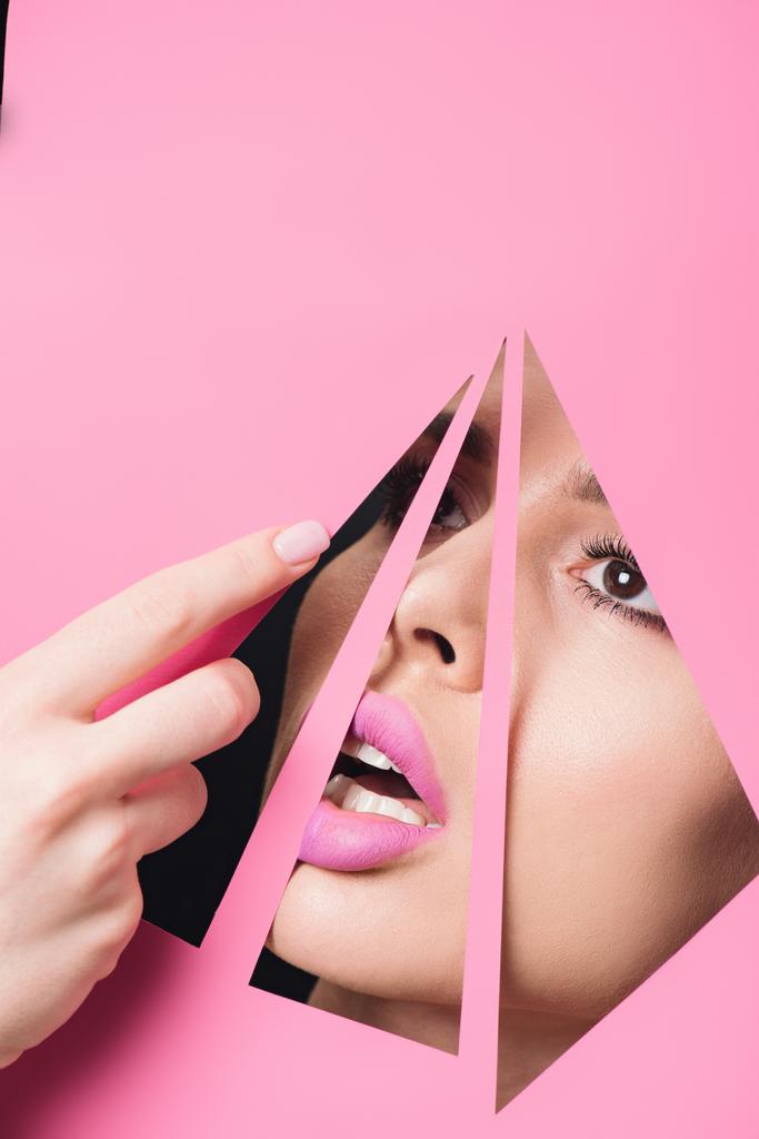 Nainen, jolla on avoin suu ja vaaleanpunaiset huulet, jotka katsovat kolmion reikien yli ja koskettavat paperia käsin mustalla taustalla
 - Valokuva, kuva