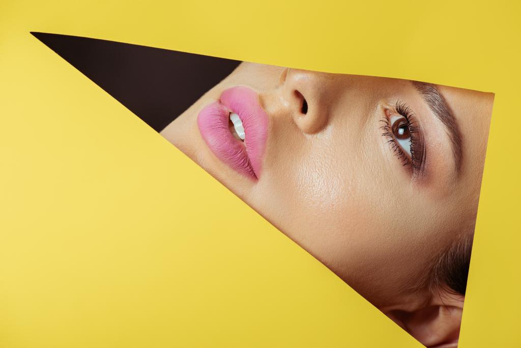 Mädchen mit rosa Lippen blickt durch ein dreieckiges Loch in gelbem Papier auf schwarz - Foto, Bild