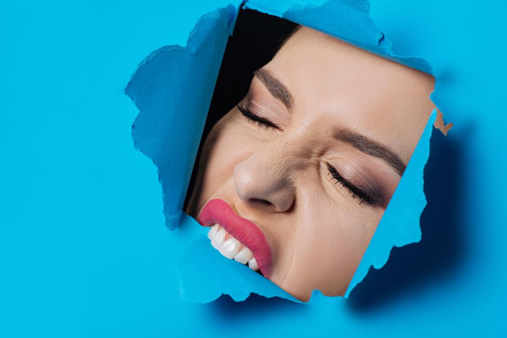 Woman with closed eyes and pink lips biting ripped blue paper on black - Photo, Image