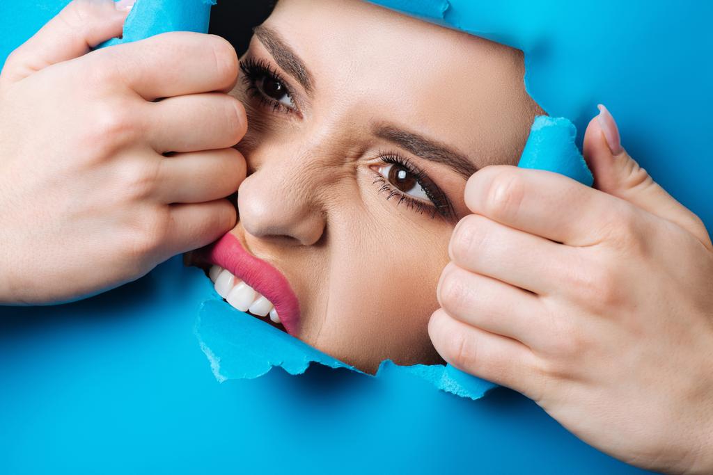Žena s růžovými rty a kouřovýma očima dotýkající se a kousající roztrhaný modrý papír přes díru - Fotografie, Obrázek