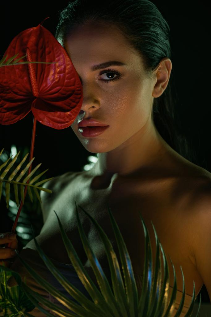 Mädchen blickt in die Kamera und bedeckt Gesicht mit rotem Anthurium in der Nähe von Blättern isoliert auf schwarz - Foto, Bild
