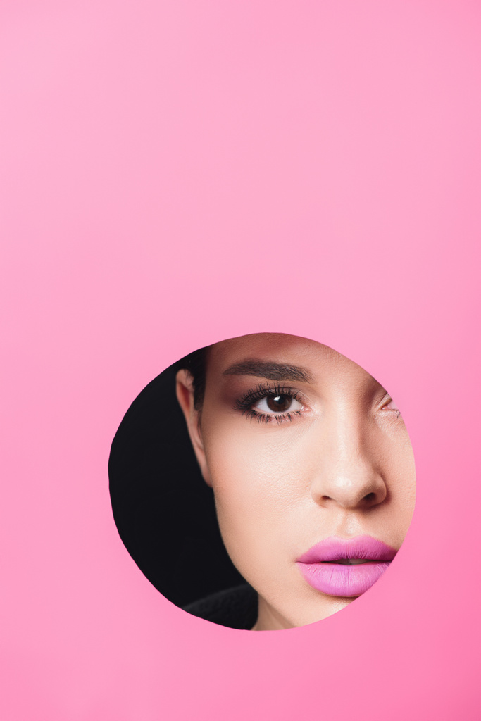 Όμορφη κοπέλα με καπνιστά μάτια και ροζ χείλη κοιτάζοντας κάμερα σε όλη την τρύπα σε χαρτί σε μαύρο - Φωτογραφία, εικόνα