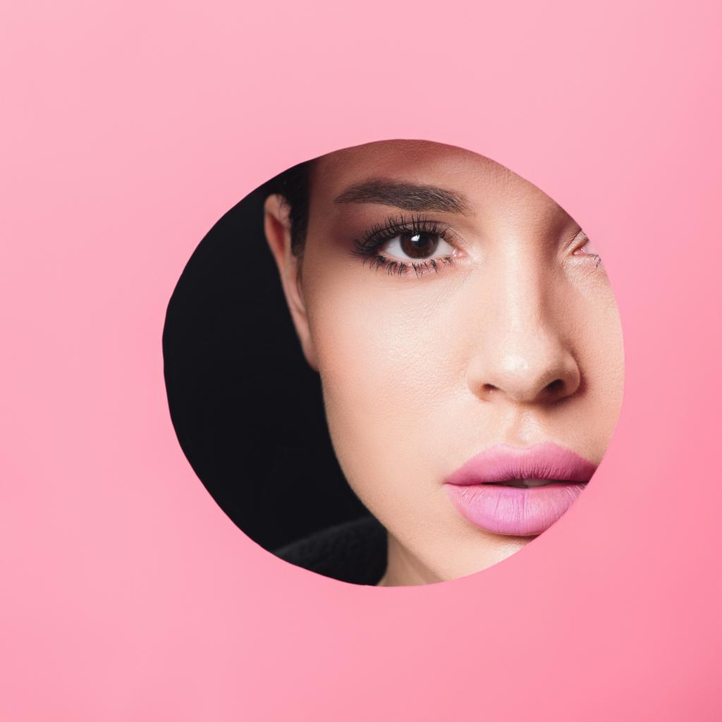 Красивая девушка с дымчатыми глазами и розовыми губами, смотрящая на камеру через отверстие в бумаге на черном фоне
 - Фото, изображение