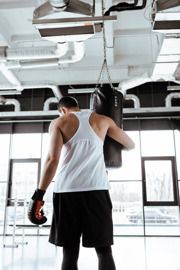 スポーツセンターに立ちながらパンチングバッグに触れるボクシンググローブのスポーツマンの後ろ姿  - 写真・画像