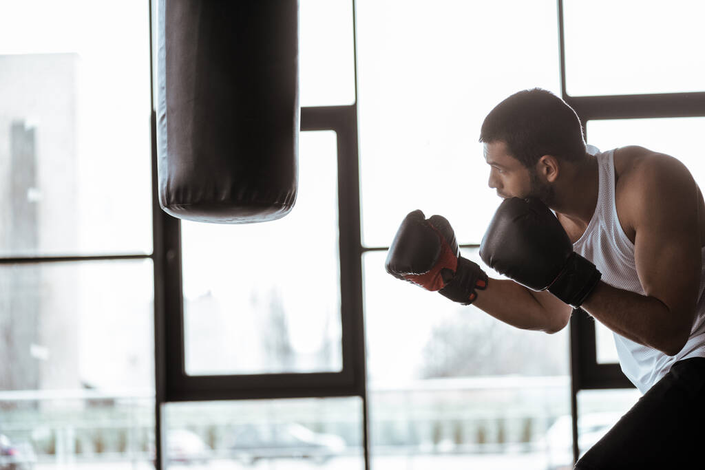 boczny widok przystojnego sportowca w rękawicach bokserskich trening z workiem bokserskim   - Zdjęcie, obraz