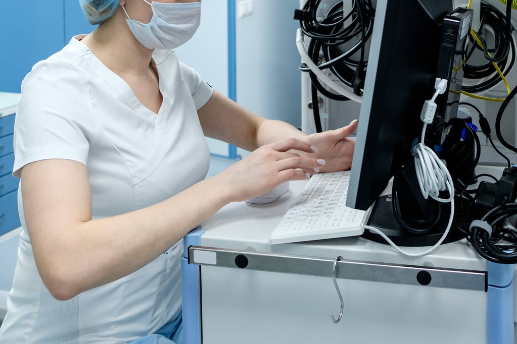 Ένας γιατρός με μάσκα και γάντια εργάζεται σε έναν υπολογιστή ιατρικού εξοπλισμού στα επείγοντα. Ανάλυση πληροφοριών σχετικά με τη λοίμωξη από τον κορωναϊό. - Φωτογραφία, εικόνα