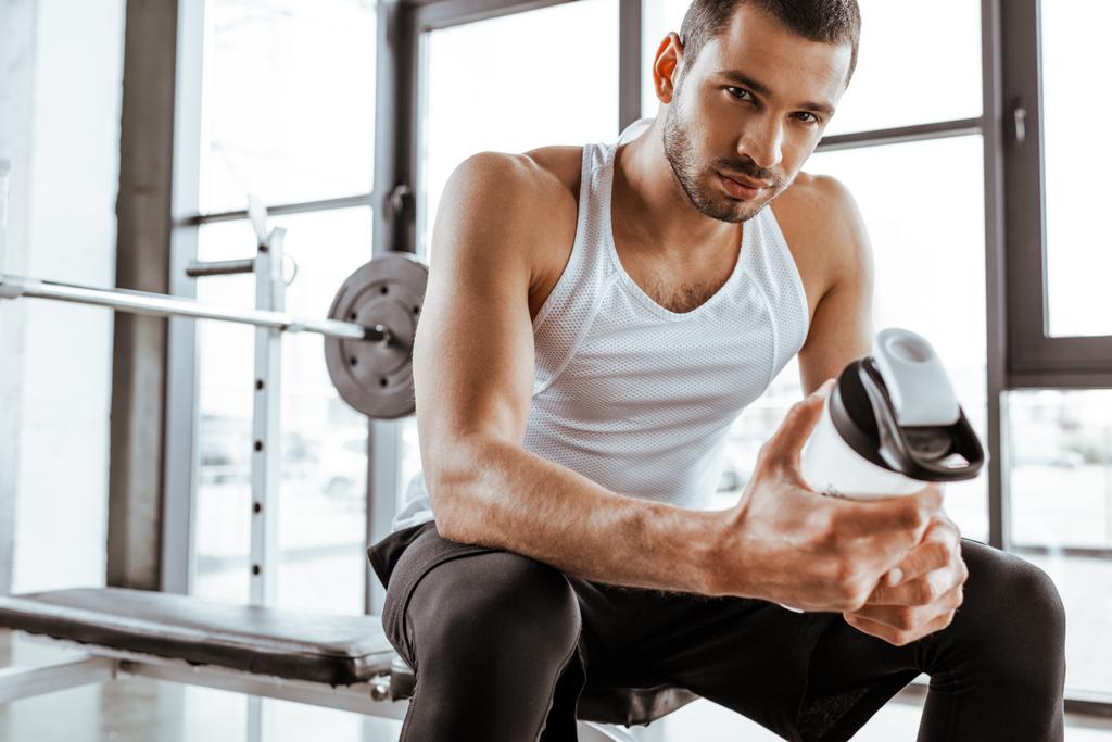 όμορφος αθλητής κρατώντας αθλητικό μπουκάλι με πρωτεΐνη milkshake και κοιτάζοντας κάμερα στο γυμναστήριο  - Φωτογραφία, εικόνα