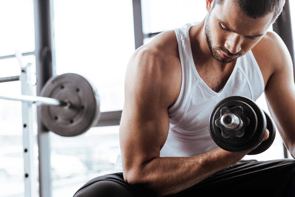 strong sportsman exercising with dumbbell while sitting in gym  - Photo, Image