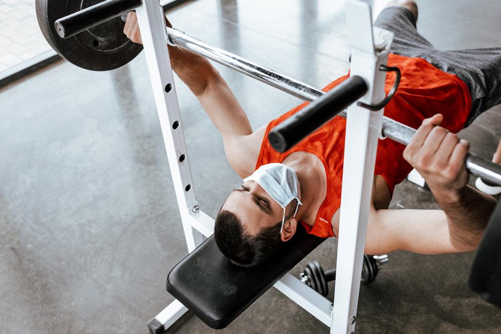 athletic sportsman in medical mask exercising with barbell in gym  - Photo, Image