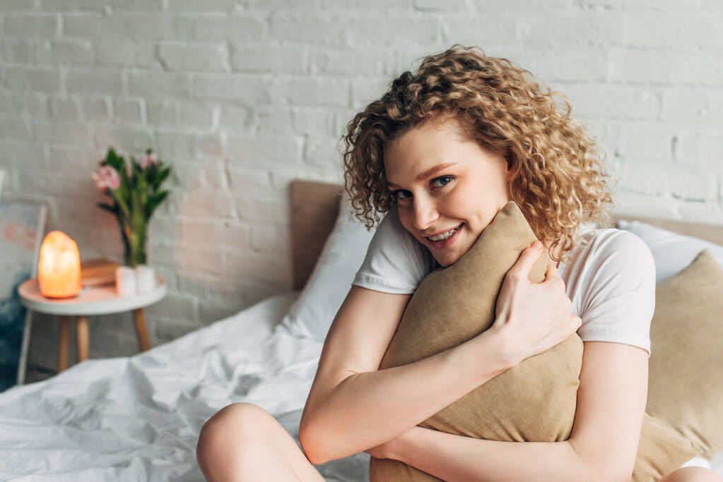 χαρούμενο όμορφο κορίτσι σε homewear αγκαλιάζει μαξιλάρι στο κρεβάτι στο άνετο υπνοδωμάτιο με λάμπα αλάτι Ιμαλαΐων - Φωτογραφία, εικόνα