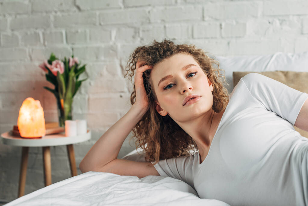 attractive curly girl in homewear lying on bed in bedroom with Himalayan salt lamp - Photo, Image