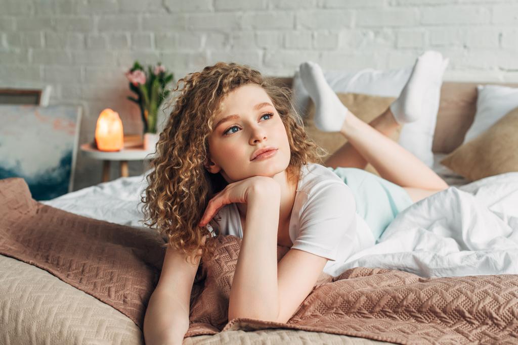 задумчивая девушка в домашней одежде лежит на кровати в спальне с гималайской соляной лампой
 - Фото, изображение
