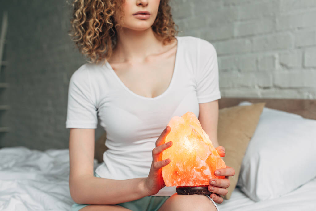 cropped view of curly woman in homewear holding Himalayan salt lamp in bedroom - Photo, Image