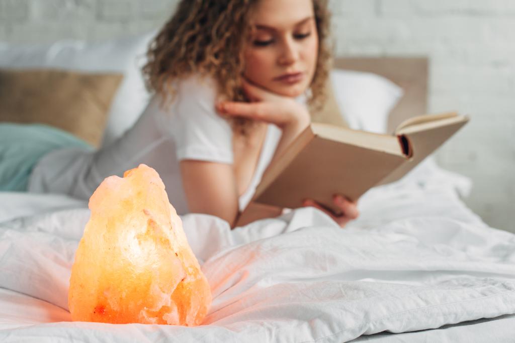Kıvırcık kadının yatakta Himalaya tuz lambasıyla kitap okuduğu seçici odak noktası  - Fotoğraf, Görsel