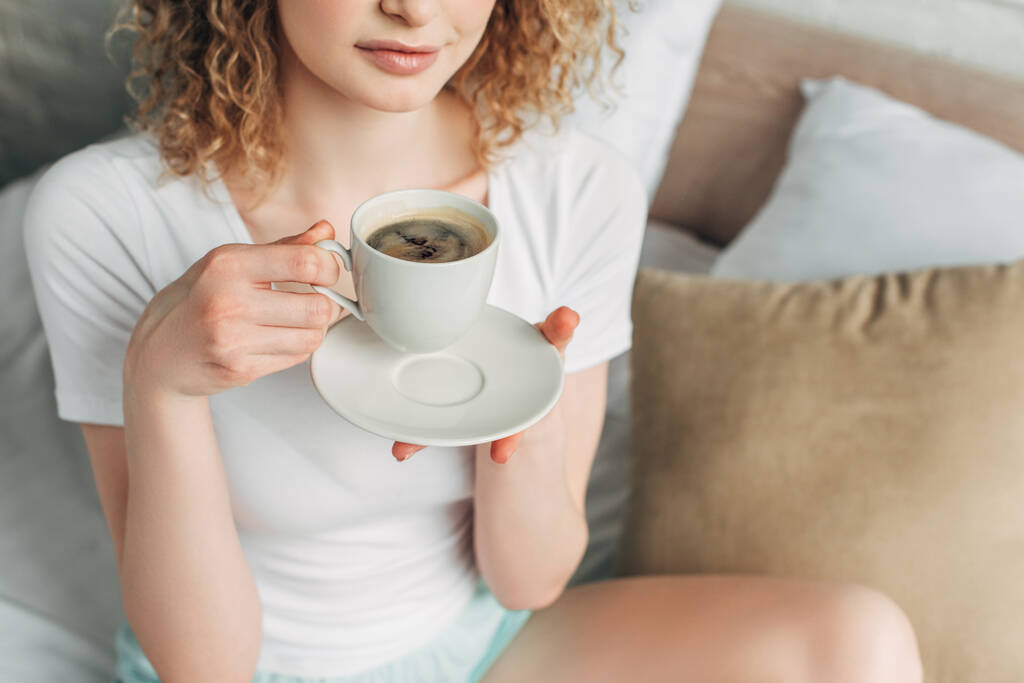İç çamaşırlı genç bir kadının sabahları elinde bir fincan kahveyle yatakta görüntüsü.  - Fotoğraf, Görsel