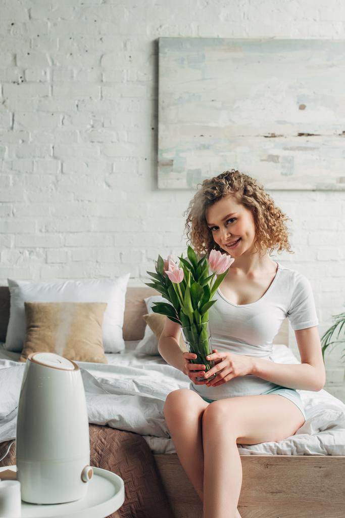chica feliz sosteniendo tulipanes mientras está sentado en el dormitorio con purificador de aire
  - Foto, imagen