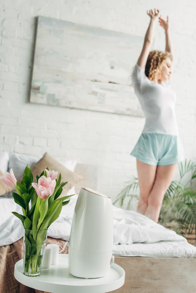 kobieta rozciągająca się w sypialni z oczyszczaczem powietrza i kwiaty, selektywne skupienie - Zdjęcie, obraz