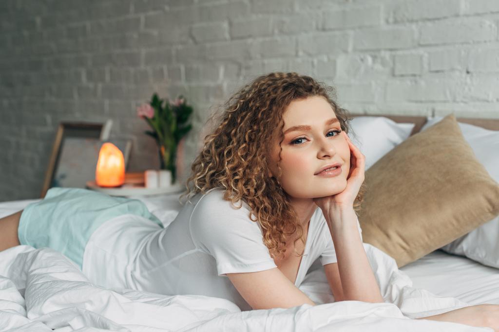 улыбающаяся девушка в домашней одежде лежит на кровати в спальне с гималайской соляной лампой
 - Фото, изображение