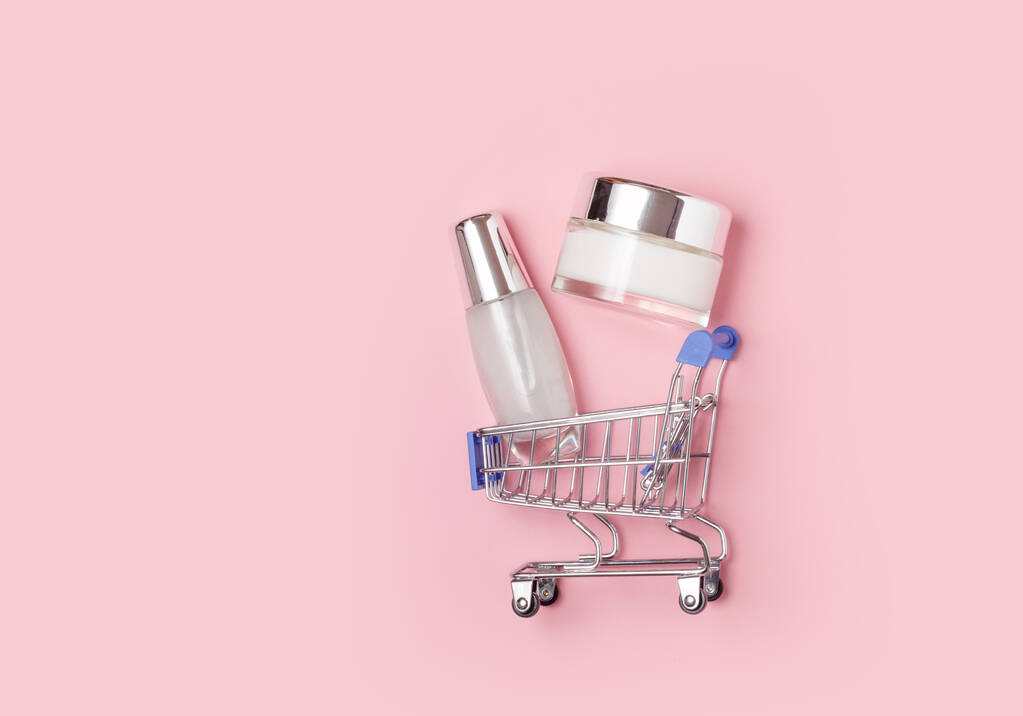 Weiße Kosmetikdosen mit Sahne liegen in einem Einkaufswagen auf rosa Hintergrund. Online-Einkauf zu Hause - Foto, Bild